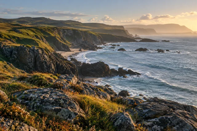 Foreground coastal rocks lead toward a curving shoreline and distant layered cliffs in late afternoon light.